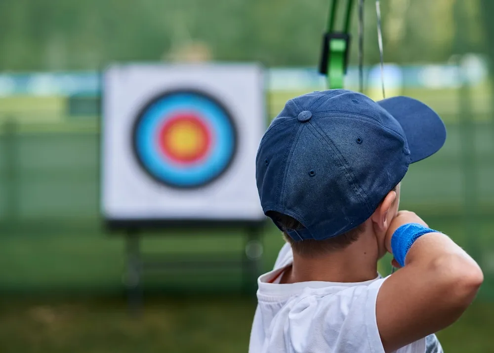 Enfant de dos pratiquant le tir à l’arc en extérieur, visant une cible colorée.